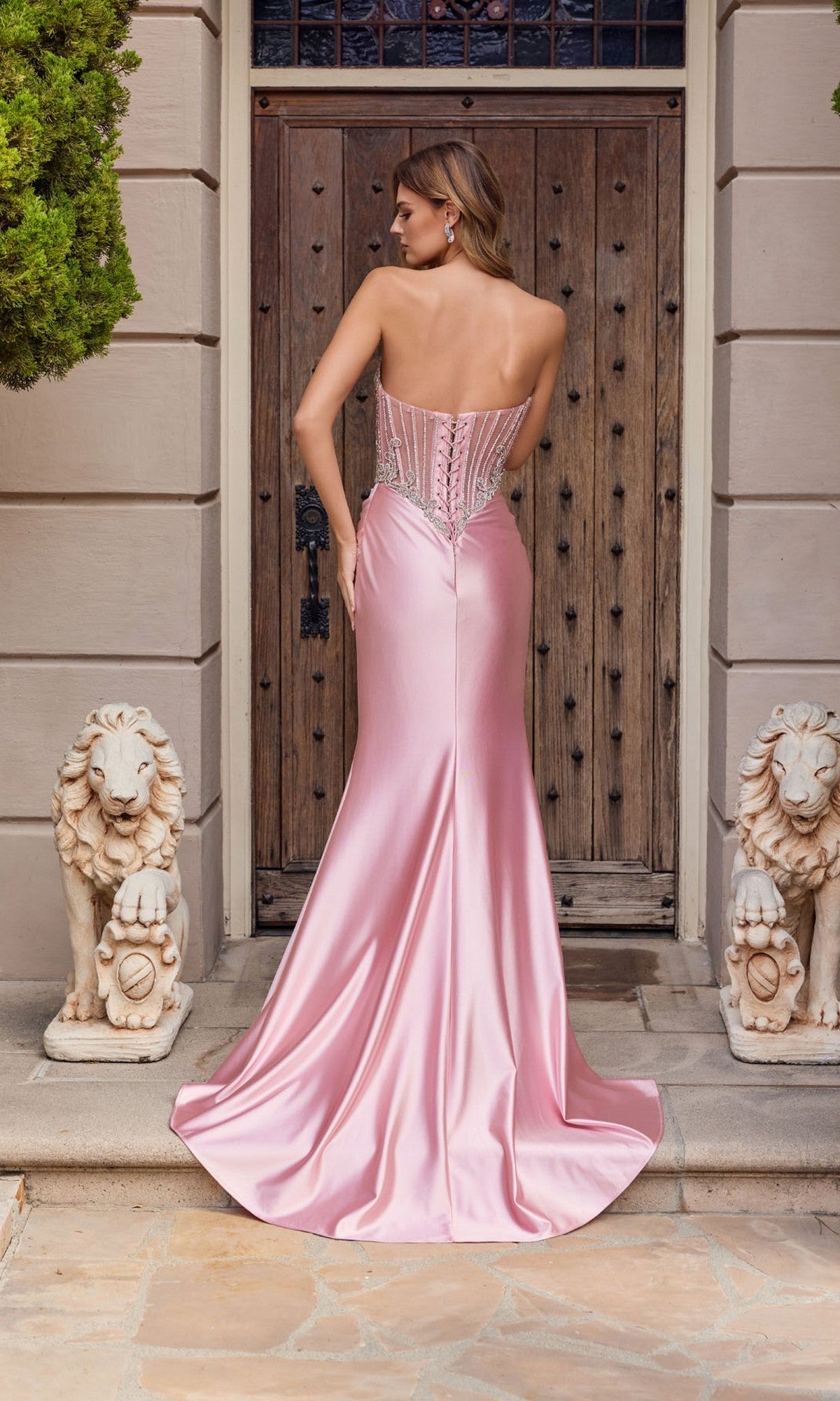 A woman in the Juliet JT2654K Long Prom Dress, featuring a strapless corset bodice and satin sheath skirt, stands with her back to the camera before a wooden door between two stone lion statues.